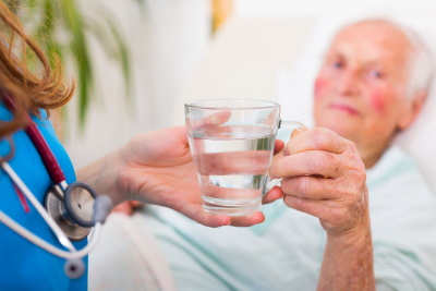 A medical professional wearing a stethoscope handing a glass of water to an elderly man who is lying in bed.