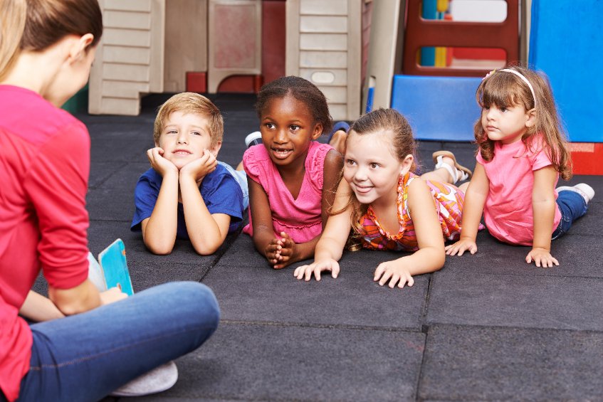 A nursery worker reading a book to 4 children lying on the ground